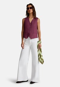 Kobieta nosząca bezrękawnik w kolorze śliwkowym z guzikami, białe spodnie o szerokich nogawkach, czarne buty, ciemne okulary przeciwsłoneczne, trzymająca zielono-paskowaną szalik na białym tle.