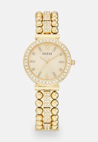 Gold watch with a round face, cream dial, crystal accents, and a beaded bracelet design. Markers at 12 and 6; logo "GUESS" displayed.
