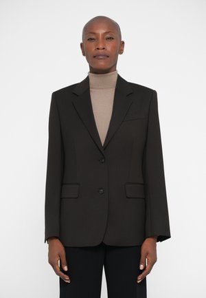 Kvinde iført en sort skræddersyet blazer over en beige turtleneck sweater, stående mod en ensfarvet hvid baggrund.