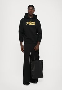 Uomo in hoodie nero Just Cavalli e pantaloni a gamba larga, che tiene una borsa a tracolla in pelle nera, in piedi contro uno sfondo bianco semplice.