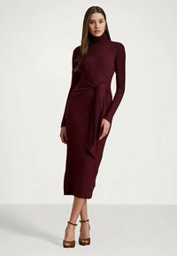 Nő hosszú ujjas, burgundi színű garbó midi ruhában, oldalsó megkötő részletekkel, keresztbe tett lábakkal pózol, barna magassarkú szandálban.