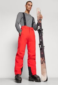 Pantalones de esquí rojos brillantes con tirantes negros ajustables, combinados con una camiseta de manga larga gris y botas de esquí negras, sosteniendo una tabla de snowboard.