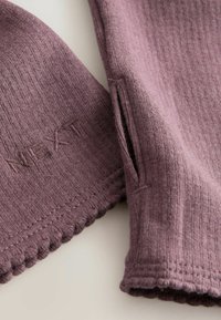 Mauve gerippter Strickstoff mit gewellten Kanten und besticktem Detail. Besitzt Schlitze als zusätzliches Designelement. Weiche Textur.