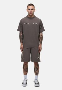 T-shirt grigia a maniche corte e pantaloni corti abbinati realizzati in tessuto morbido, con logo stampato in bianco. Il modello indossa sneakers nere con calzini bianchi.