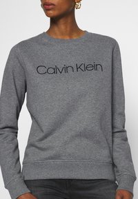 Sudadera gris con el logotipo "Calvin Klein" en negro, cuello redondo, puños y bajo de canalé, tela suave y corte relajado.