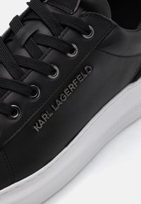 Black leather sneaker with round toe, flat black laces, silver eyelets, and white rubber sole. Features embossed branding on the side.