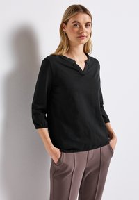 Zwarte blouse met lange mouwen en een V-halslijn, met elastische manchetten, gecombineerd met lichtbruine, op maat gemaakte broek met zijzakken.