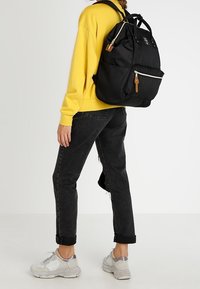 Sac à dos noir avec des fermetures éclair et des accents en cuir marron, porté sur un sweat-shirt jaune, accompagné d'un jean noir retroussé et de baskets grises.