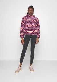 Polo i lila med geometriska mönster i rosa och orange. Fleece-material, med huva. Svarta tights och ljusa träningsskor kompletterar outfiten.