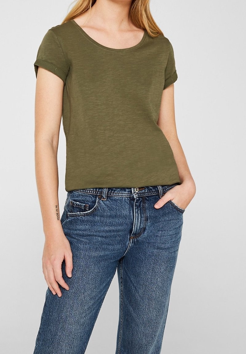 T-shirt vert olive à manches courtes roulées, col rond et coupe décontractée, porté avec un jean taille haute en denim bleu.