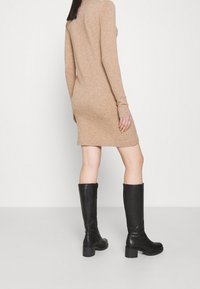 Robe beige en maille à manches longues avec des poignets côtelés, associée à des bottes en cuir noir montantes à talon épais et surface texturée.