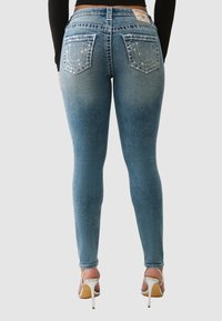 Jean denim bleu skinny taille haute, délavé, poches arrière ornées et patch logo sur la ceinture. Porté avec des talons hauts beige.