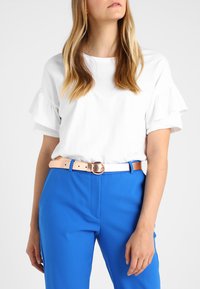 White short-sleeve top with ruffled sleeves, paired with bright blue trousers and a tan belt featuring a round metal buckle.