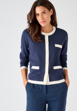 Vrouw met bruin haar draagt een marineblauwe cardigan met witte bies en knopen, gecombineerd met marineblauwe broek met krijtstrepen, handen in zakken, neutrale achtergrond.