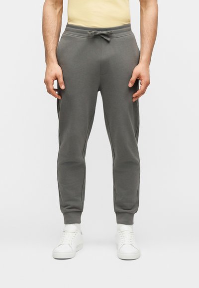 Homme portant un pantalon de jogging gris avec taille à cordon, associé à des baskets blanches, debout devant un fond uni.