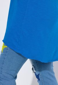 Blaues Hemd mit abgerundetem Saum, kombiniert mit hellblauen Jeans, die gelbe und blaue Sternaufnäher sowie Abrieb an den Knien aufweisen.