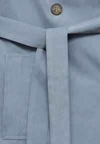 Svetlofialová látka s saténovým povrchom, s opaskom zviazaným v páse, gombíkom a obdĺžnikovou vreckom na ľavej strane.