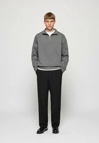 Pelēks vilnas džemperis ar puszipu apģērbts virs baltas T-krekla, kombinēts ar melnām biksēm un melniem apaviem.