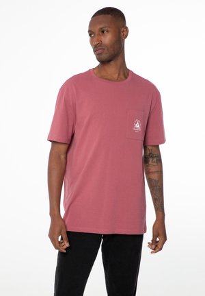 T-Shirt mit kurzen Ärmeln und Rundhalsausschnitt in staubrosa, mit einer linken Brusttasche, auf der ein weißes geometrisches Logo zu sehen ist. Hergestellt aus weichem Baumwollstoff.