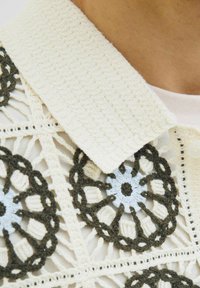 Kvačkana bluzica s kremastim ovratnikom, ki ima zapletene cvetlične motive v temno zeleni in svetlo modri niti na teksturirani podlagi.