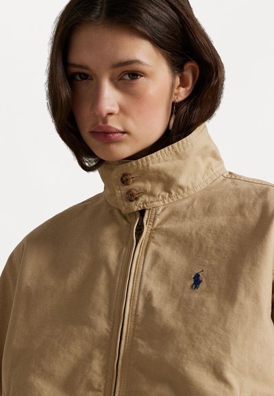 Beige Jacke mit Stehkragen, zwei Knopfakzenten und einem blauen Logo auf der linken Seite. Das Material wirkt strukturiert und robust.
