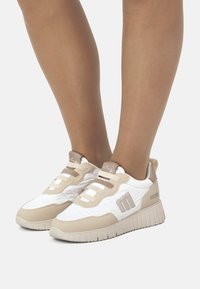 Las zapatillas deportivas en blanco y beige cuentan con una parte superior texturizada, un cuello acolchado y una suela de goma con agarres prominentes.