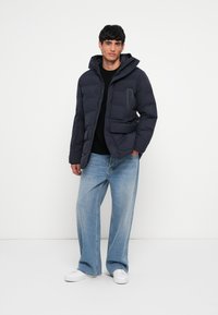 Navy puffer jacket with a zippered front, adjustable hood, and two front pockets; worn over a black shirt and light blue jeans.