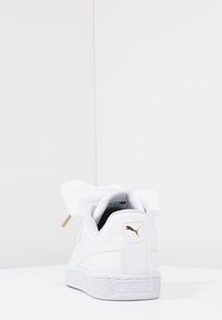 Bílá teniska z hladké kůže s texturovanou gumovou podrážkou, velkými tkaničkami ve tvaru látkového mašle a zlatým logem Puma na patě.