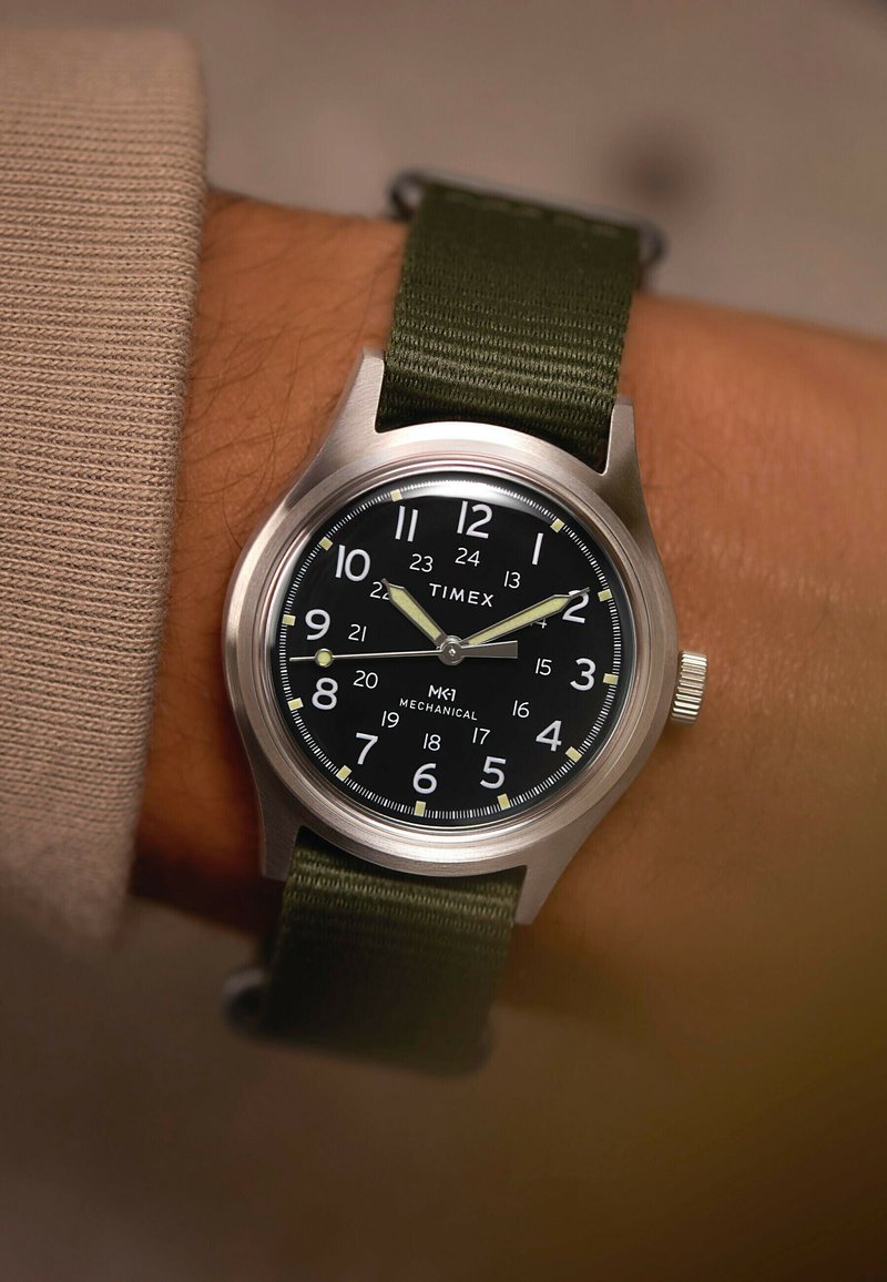 Reloj mecánico Timex MK1 con esfera negra, números blancos y correa de tela verde en una muñeca mostrando las 10:11:44.