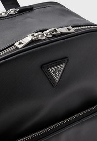 Sac en tissu noir texturé avec fermetures éclair argentées, un badge logo triangulaire et un design élégant. Présente une fermeture éclair proéminente et des poignées.