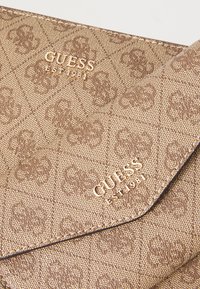 Pochette texturée brune avec un motif matelassé, arborant le logo doré en relief "GUESS EST 1981" sur le devant. Design élégant de fermeture à rabat.