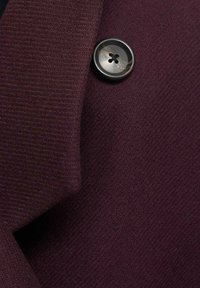Bordeaux stof med en tekstureret vævning, prydet med en mørk knap med fire huller. Materialet ser blødt og let tykkere ud.