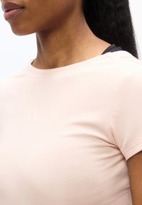 Κοντινό πλάνο προσώπου ατόμου που φοράει ροζ ανοιχτό κοντομάνικο πουκάμισο με διακριτικό ανάγλυφο λογότυπο στο στήθος και μαύρα μαλλιά που είναι μαζεμένα πίσω.