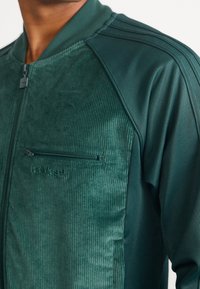 Grön zip-jacka med en kordtyptextur framtill, slätt tyg på ärmarna och en zip-fickan på bröstet.