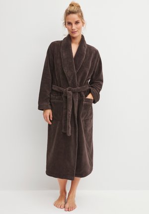 Vrouw met blond haar in een knot, gekleed in een lange, donkerbruine, zachte badjas met een gestrikte ceintuur en twee voorzakken, blootsvoets staand tegen een witte achtergrond.