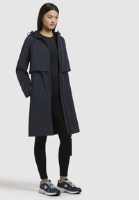 Manteau imperméable bleu marine avec capuche, fermeture éclair sur le devant et grandes poches ; associé à des leggings noirs et des baskets pour un look décontracté.