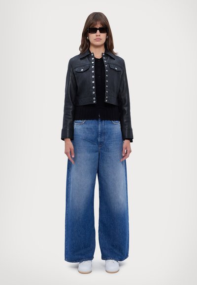 Woman wearing black cropped leather jacket, black shirt, wide-leg blue jeans, white sneakers, and rectangular black sunglasses, standing against white background.