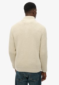 Dzianinowy kremowy sweter z wysokim kołnierzem, prążkowanymi mankietami i dołem, teksturowanym wzorem oraz luźnym krojem, w połączeniu z niebieskimi dżinsami.