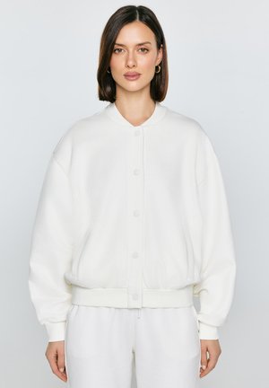 Moteris su tiesiais tamsiais plaukais dėvi baltą užsegamą bomberio stiliaus striukę ir baltas kelnes, stovinti priešais šviesią, vienalytę foną.