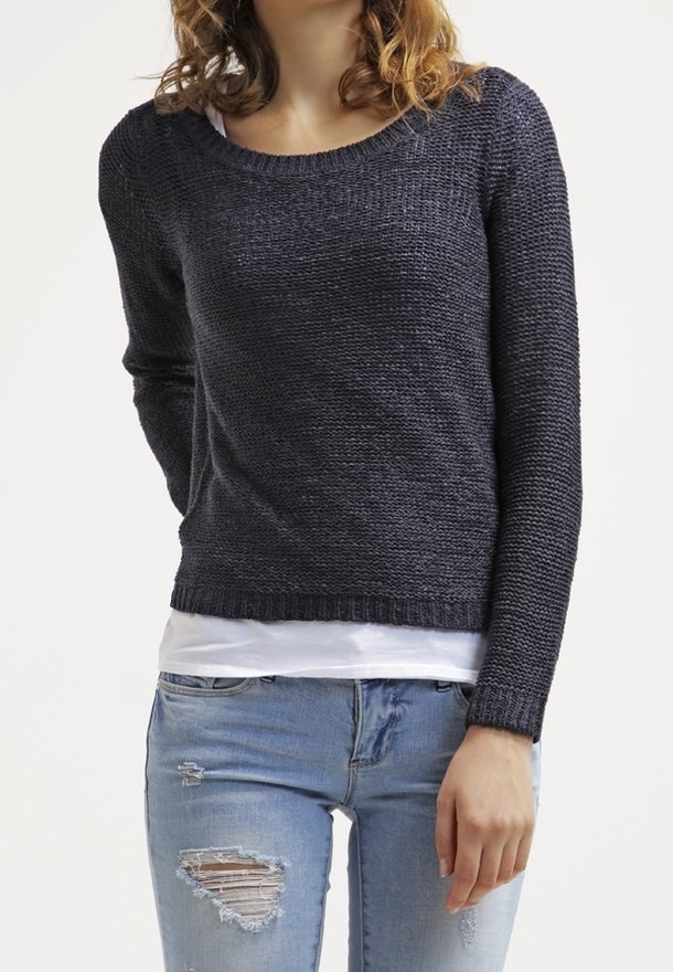 Femme portant un pull en tricot foncé par-dessus une chemise blanche et un jean bleu clair déchiré, debout devant un fond blanc uni.