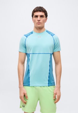 Muž stojaci čelom vpred, oblečený v svetlomodrom športovom tričku s tmavomodrými švami a svetlozelených šortkách s čiernym logom.