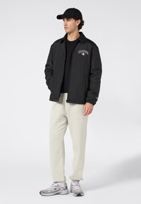Jongeman in zwarte pet en jas, crèmekleurige trainingsbroek en grijze sneakers, staande met handen in zijn zakken op een effen achtergrond.