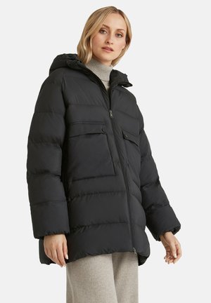 Femme portant un manteau d'hiver matelassé noir avec des poches avant et une capuche, associé à un col roulé gris clair et un pantalon, debout sur fond blanc.