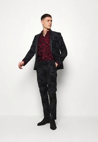 Schwarz gemusterter Anzug mit einem dunklen Blumenmuster, kombiniert mit einem weinrot strukturierten Hemd. Das Model steht selbstbewusst da und trägt schwarze Schuhe.