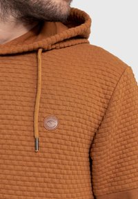 Haut à capuche en tissu texturé brun avec un motif tissé doux. Il dispose d'une capuche avec cordon de serrage et d'un patch logo rond sur la poitrine.
