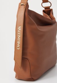 Ruda odinė pečių rankinė su tekstūruota paviršiumi, ant dirželio esantis auksinis "Valentino" logotipas ir laisvos formos dizainas.