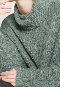 Pull en tricot vert avec un col roulé haut à côtes et une coupe décontractée. Tissu texturé avec des coutures visibles et des manches longues et larges.