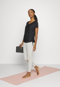 Zwarte V-hals top met korte mouwen en zijbandjes, gecombineerd met lichtgrijze broek met elastische boorden, gedragen op een roze yogamat.