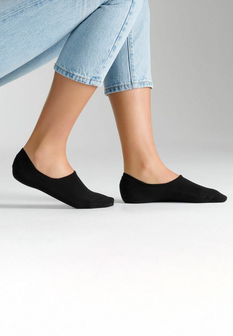 Schwarze Knöchelsocken mit einer weichen, glatten Textur und minimalistischem Design, kombiniert mit hellblauen, hochgekrempelten Jeans, die einen tiefen Ausschnitt zeigen.