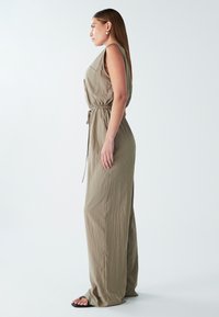 Combinaison en khaki avec une coupe ample, design sans manches, taille à cordon de serrage et texture subtile. Portée avec des sandales noires, sur fond uni.